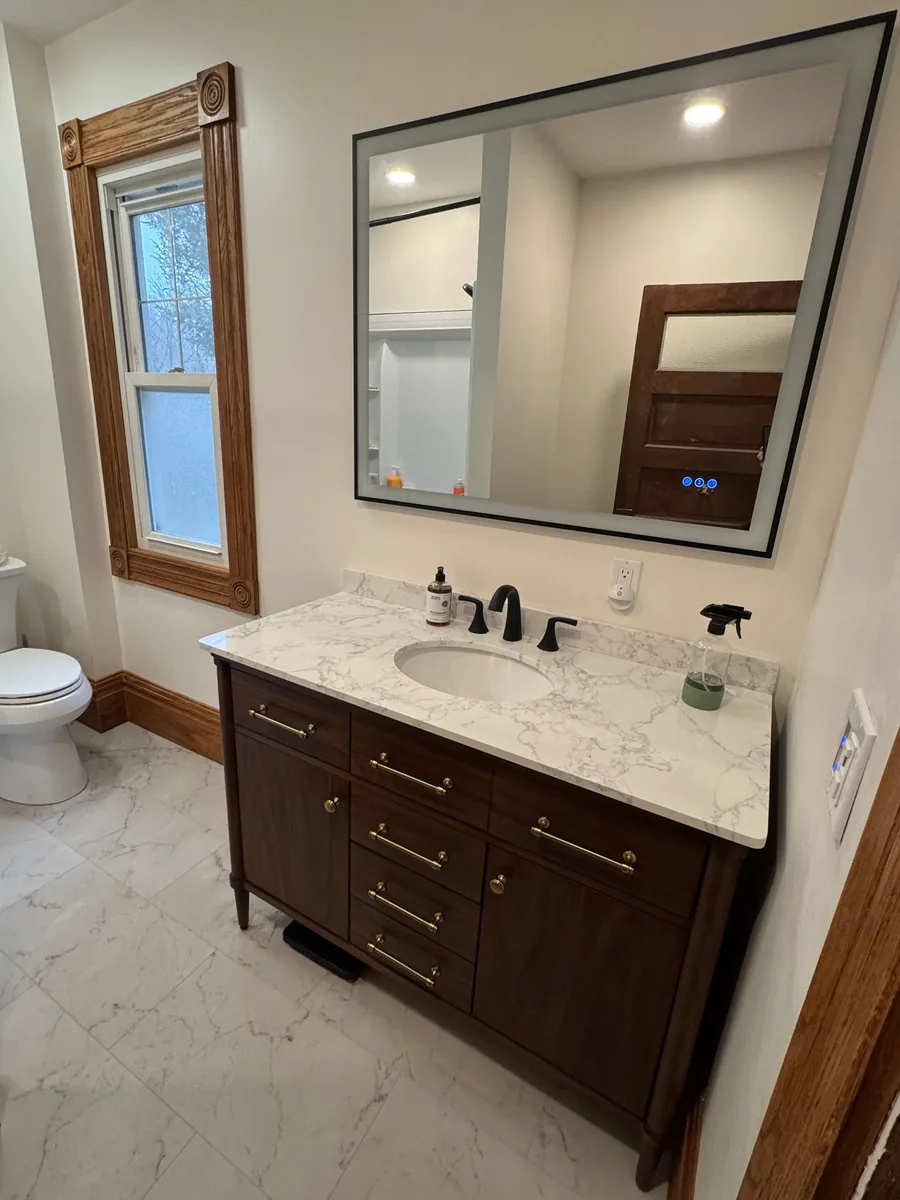 Completed Frontline bathroom project with marble-look floor, dark wood vanity, large mirror, and restored wood window trim