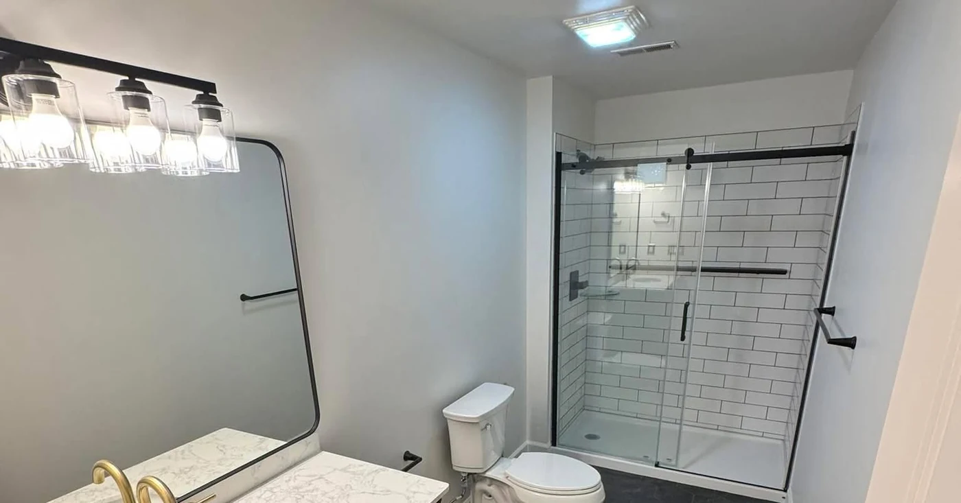 Completed Frontline bathroom remodel showing the full room, wood vanity, dark hexagonal floor tile, toilet, and black-framed tiled shower