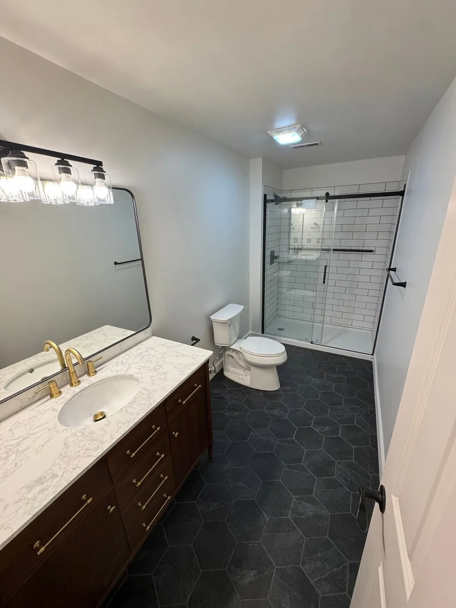 Completed Frontline bathroom remodel with wood vanity, marble-look countertop, black hexagonal floor tile, and tiled walk-in shower